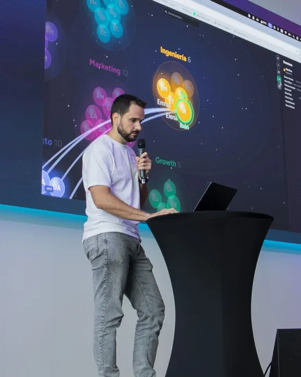 Ibán Ríos presenting on stage, microphone in hand, in front of a giant screen